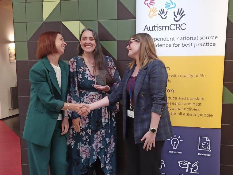 A woman in a green suit shakes hands with another woman, wearing a patterned blazer, while another woman stands between them smiling and holding an award.