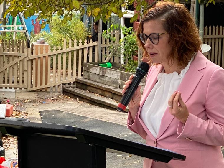 A woman speaks into a microphone, behind her is a playground.