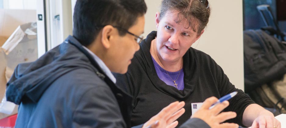 Two people sitting next to each other at a table. One person is talking while the other is listening. They are working together on an activity.