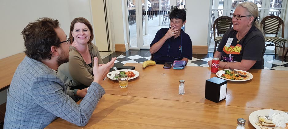 Four people sit at a dining table eating lunch. They are having a conversation and smiling.