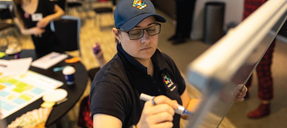 A person is in the foreground wearing a baseball cap. They are writing on a whiteboard. There are three other people in the background discussing ideas.