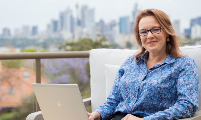 Kathleen sits in front of a laptop on a sunny balcony.