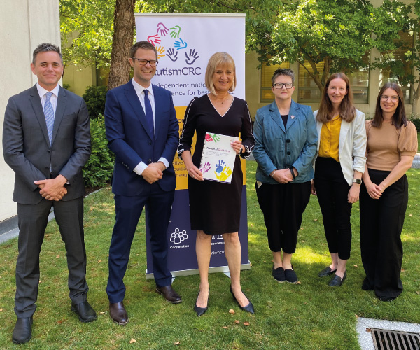 The Guideline Development Group and Justine Elliot standing in front of an Autism CRC banner. Justine Elliot holds a copy of the Guideline.