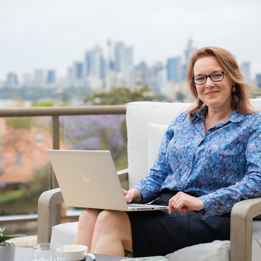 Kathleen sits in front of a laptop on a sunny balcony.