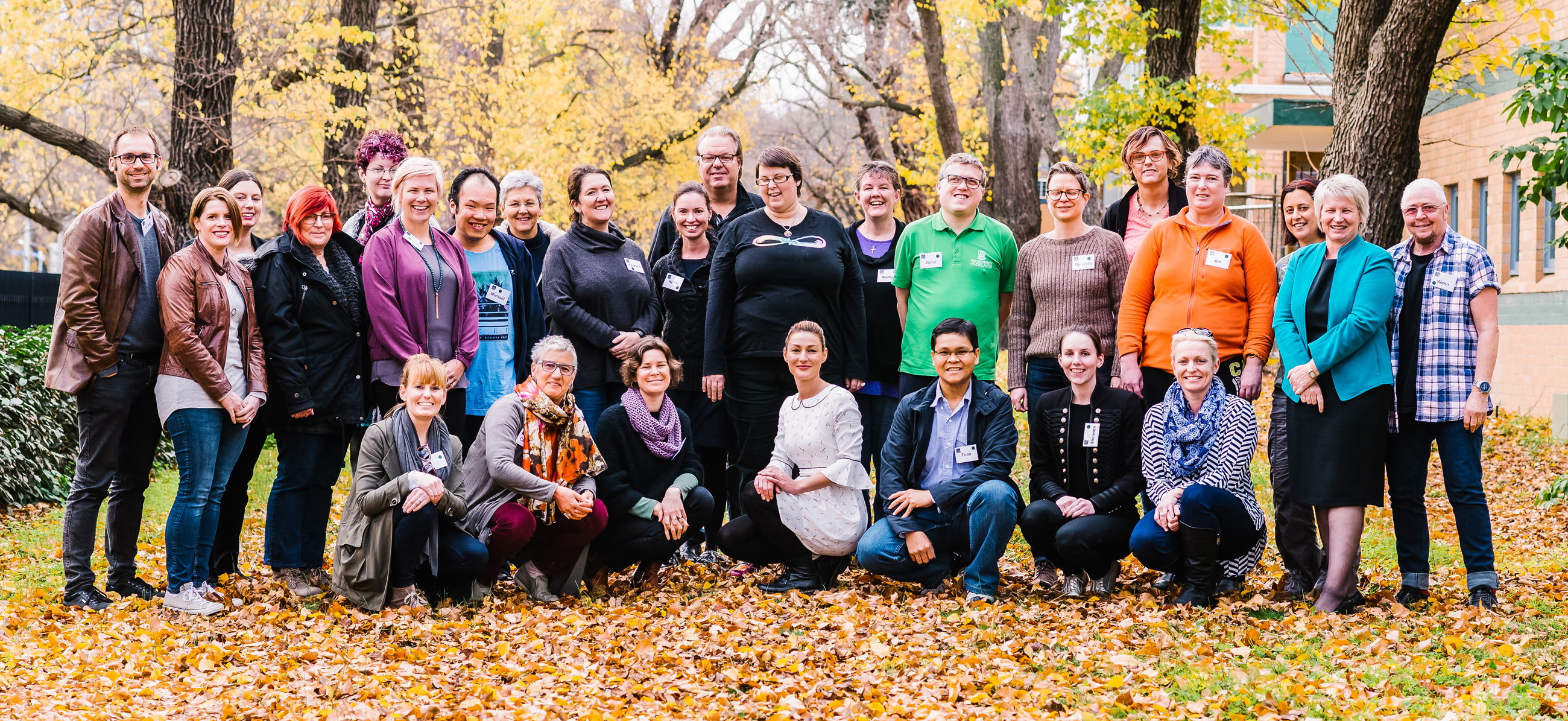 A large group of people are gathered togethered, in the midst of fallen yellow leaves.
