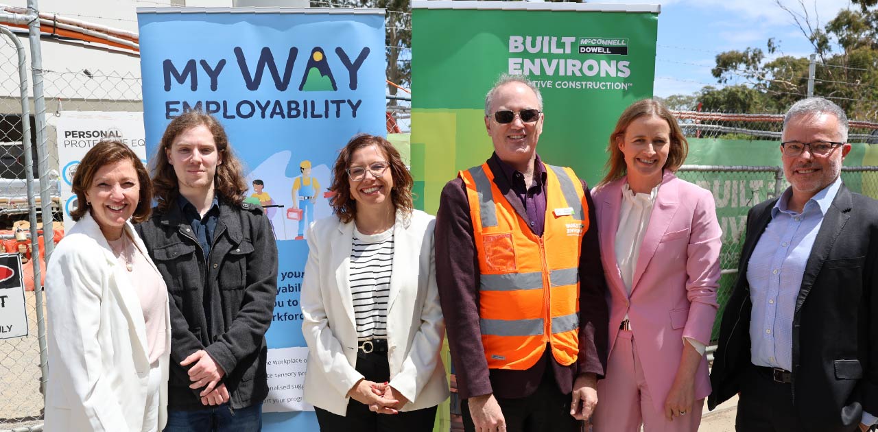 A group of people stand together in front of a construction fence and a promotional banner for myWAY Employability